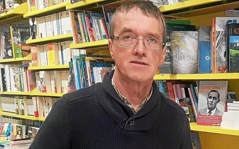 L'écrivain Roland Cotty habillé d'un pull noir devant les rayons de la Maison de la presse de Châteauneuf-du-Faou
